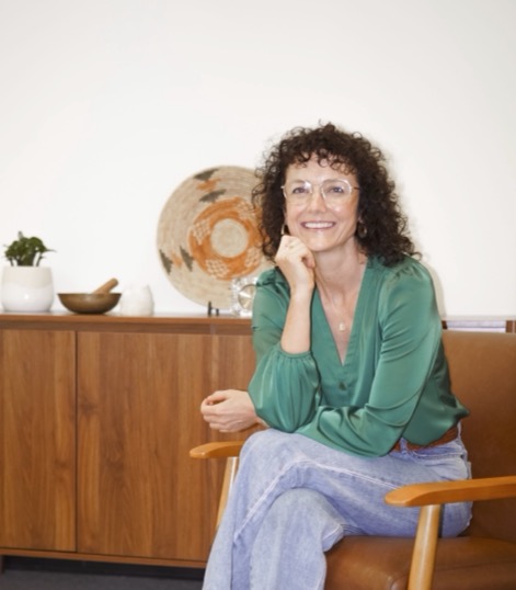 Crop this exact photo so the bottom of the frame cuts off just above the ankles/shoes, removing the sandals entirely. Keep everything else identical — the woman in the green blouse sitting in the wooden chair, the warm wood sideboard behind her, the woven basket on the wall, the rug. Do not alter colors, lighting, or composition in any other way.